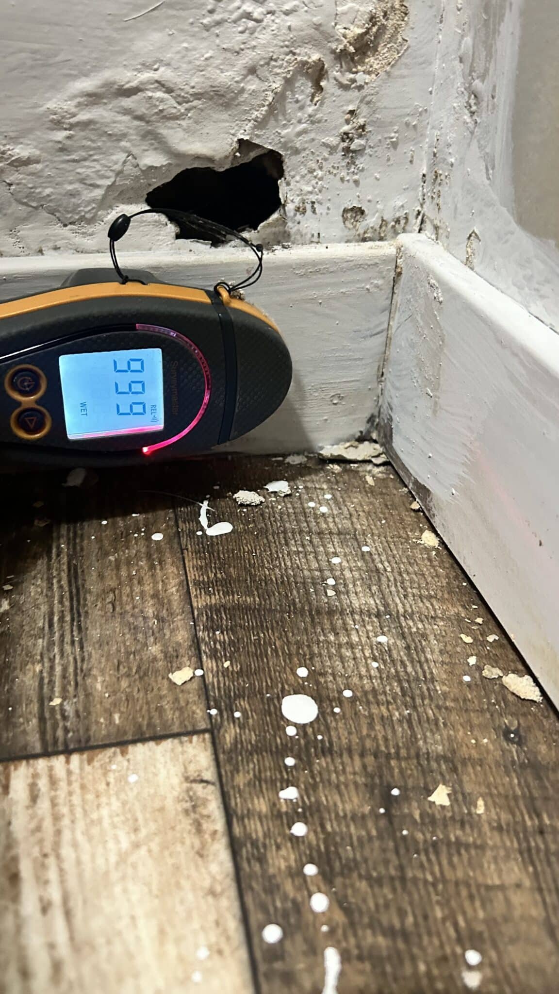 Moisture meter used beside a damaged baseboard and exposed flooring